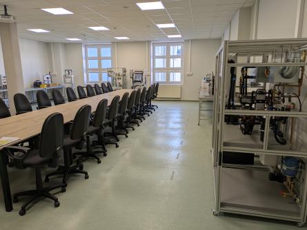 Sala laboratoryjna z długim stołem i stanowiskami komputerowymi oraz urządzeniami technicznymi