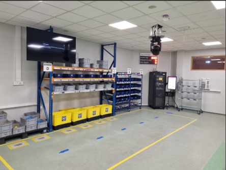 Stanowisko dydaktyczne symulujące magazyn z regałami, pojemnikami i systemem Pick-by-Light, wyposażone w monitory i urządzenia automatyki.