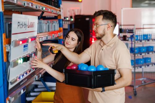 Dwie osoby obsługują system magazynowy z panelem Pick-by-Light, korzystając z czujnika i pojemnika z elementami