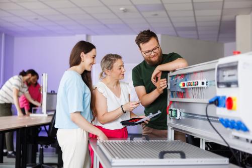 Grupa osób pracuje wspólnie przy stanowisku laboratoryjnym z urządzeniem elektrycznym; analizują okablowanie i elementy sterujące.